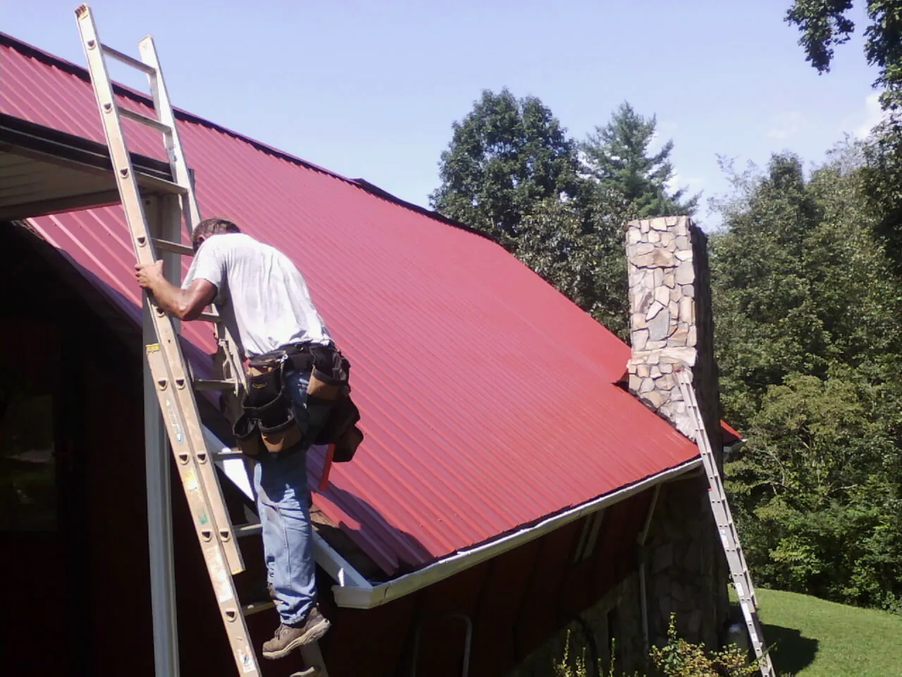Roofer on a ladder beside a red metal roof and stone chimney — Soffit & Fascia Repair by Roofing Brothers in Wilberforce